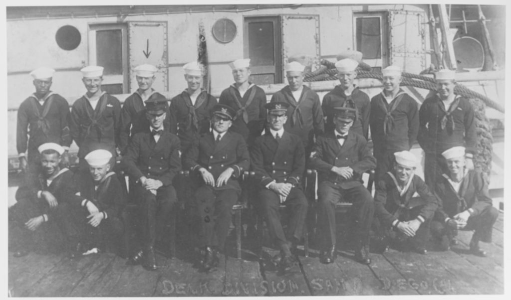 (AT-54) Deck Division posed beside the ship, at San Diego, California, circa early 1921. Seated in chairs are (from left to right): Chief Carpenter's Mate John Wesley Powell; Boatswain Harvey H. Reinbold, Executive Officer; Boatswain Roy E. Hoffses, Ordnance Officer; and Chief Boatswain's Mate Elias Melvin Zimmerman. The Sailor marked by an arrow (top row) may be Seaman 1st Class W.P. Burbage. Courtesy of W.P. Burbage, 1970. U.S. Naval History and Heritage Command Photograph. (# NH 71507)