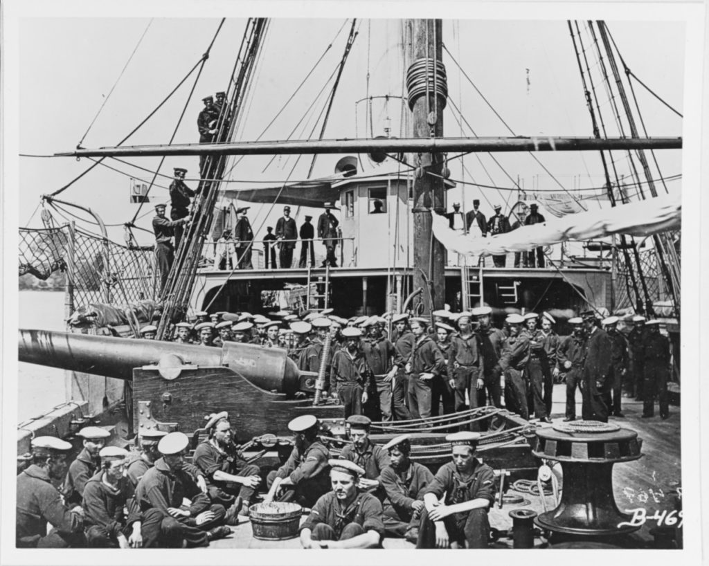 Ship's officers and crew on the foredeck, 1864-65. Photographed by Matthew Brady. Note 100-pounder Parrott rifled gun on a pivot carriage; men wearing white flat hats with the ship's name on the hat ribbon; foremast and yard; anti-boarding netting; and capstan. The original negative is # 111-B-469 in the National Archives. U.S. Naval History and Heritage Command Photograph.